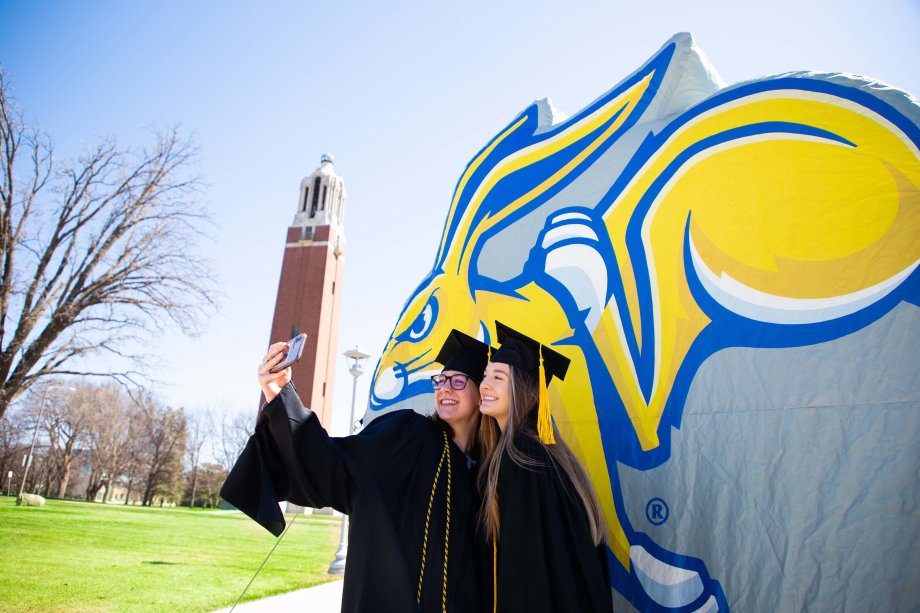 Two graduates in regalia taking a photo with a Jackrabbit inflatable and the campanile in the background