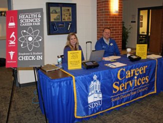 Two career professionals sitting at a table with a table drape that says Career Services, South Dakota State University at the health sciences career fair.